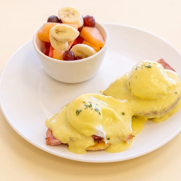 Eggs Benedict with hollandaise sauce and a side of fresh fruit salad on a white plate.