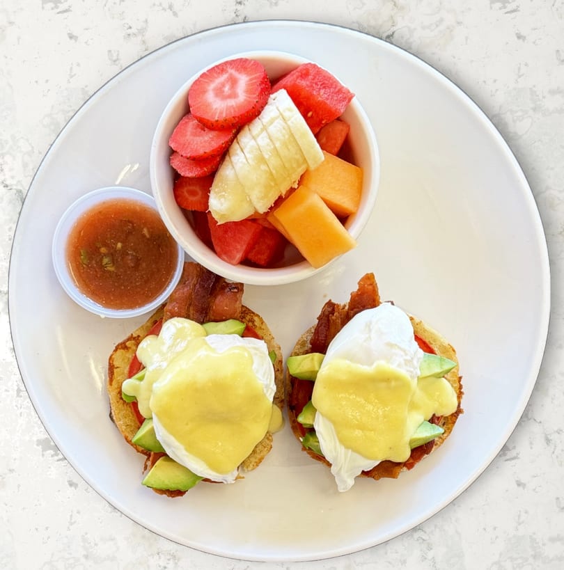 Two eggs Benedict with avocado and bacon, served with a side of fruit and salsa.