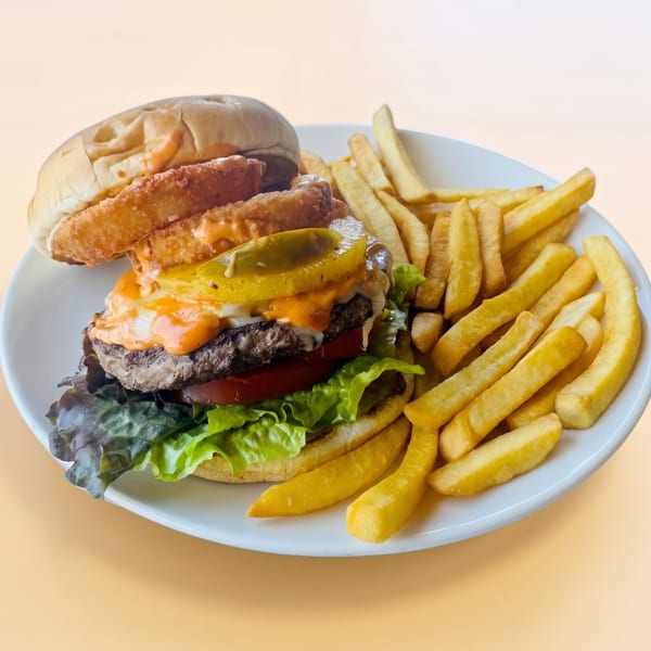 A cheeseburger with onion rings, jalapeños, lettuce, tomato, and sauce, served with golden fries on a white plate.