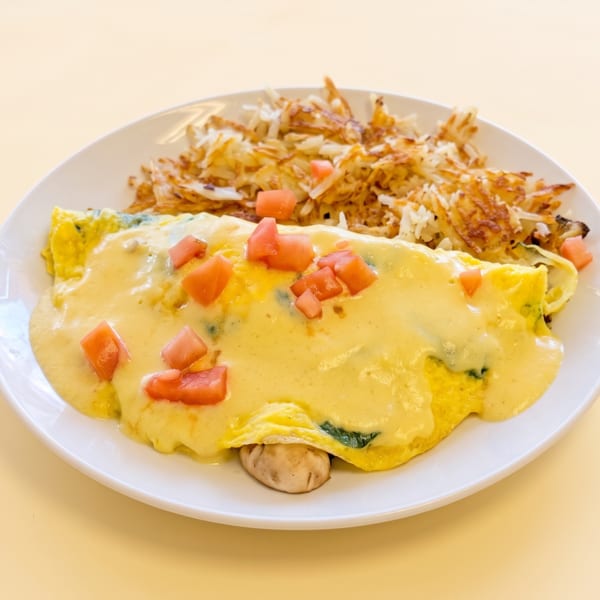 Omelet topped with cheese sauce and diced tomatoes, served with golden hash browns on a white plate.