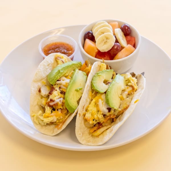 Two breakfast tacos with eggs, cheese, bacon, avocado, fruit salad, and salsa.