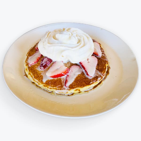 Golden pancakes topped with sliced strawberries, whipped cream, and a sweet drizzle on a white plate.
