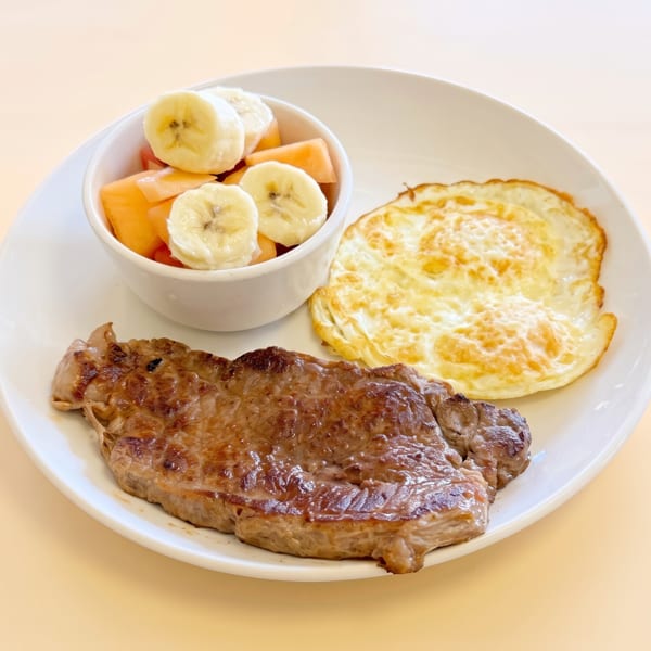 A white plate holds a steak, two fried eggs, and a bowl of fruit salad with bananas and melon.