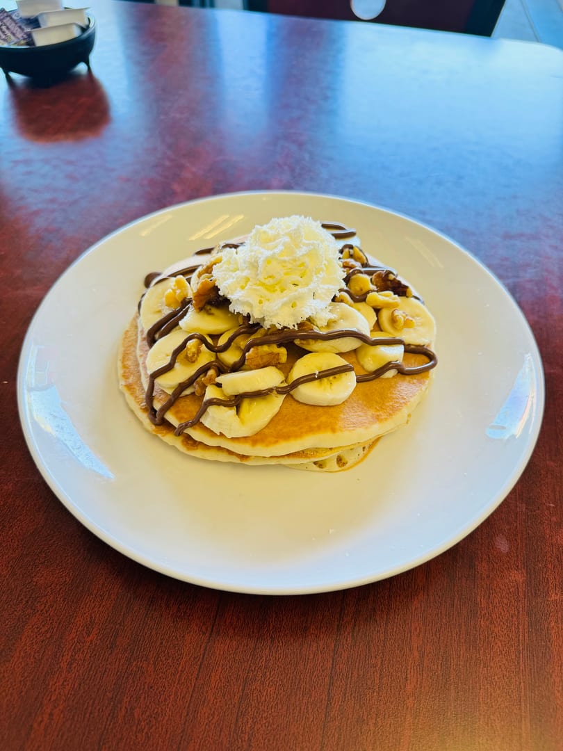 Pancakes topped with sliced bananas, walnuts, chocolate drizzle, and whipped cream on a white plate.