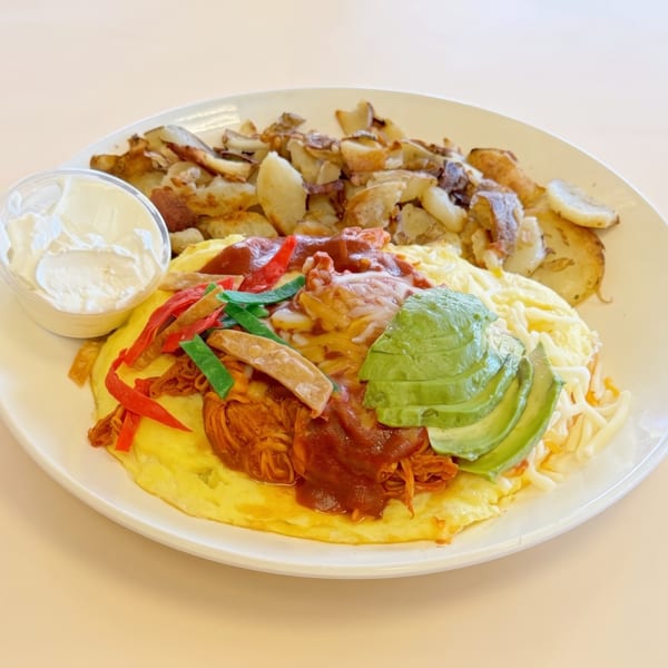 Omelet topped with shredded meat, avocado, and sauce, served with home fries and sour cream.