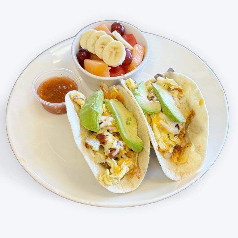 Two breakfast tacos with avocado, scrambled eggs, cheese, and bacon, served with fruit and salsa.