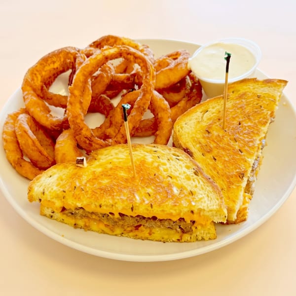 Grilled sandwich with melted cheese and a side of crispy onion rings and dipping sauce on a white plate.