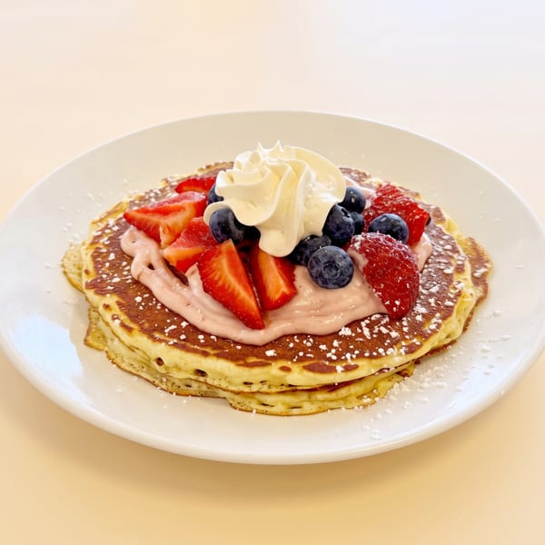Stack of pancakes with pink cream, strawberries, blueberries, whipped cream, and powdered sugar on a white plate.